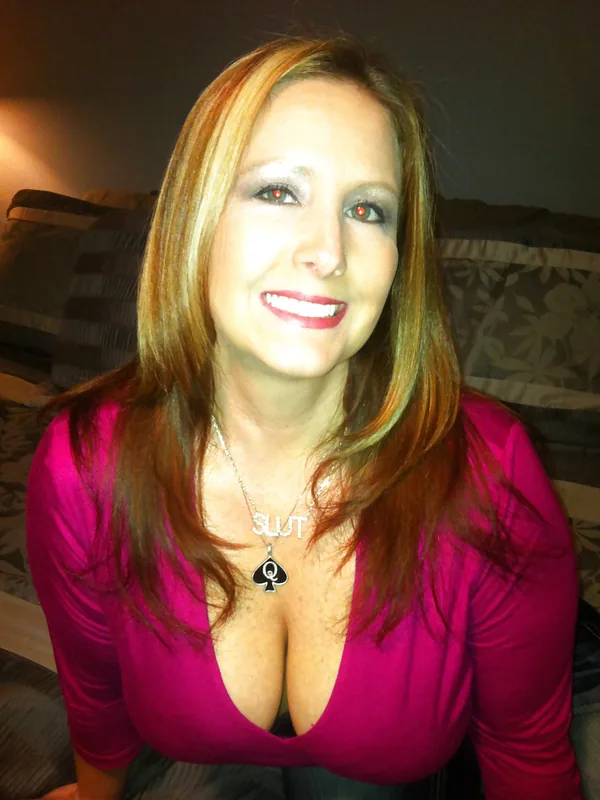 Femme aux cheveux châtain clair en haut rose, collier 'SLUT', souriante, assise sur un canapé.