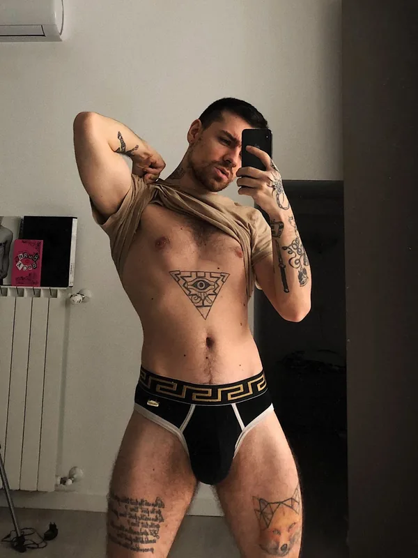Homme tatoué prenant un selfie dans un miroir, torse nu, portant des sous-vêtements noirs et un t-shirt relevé.