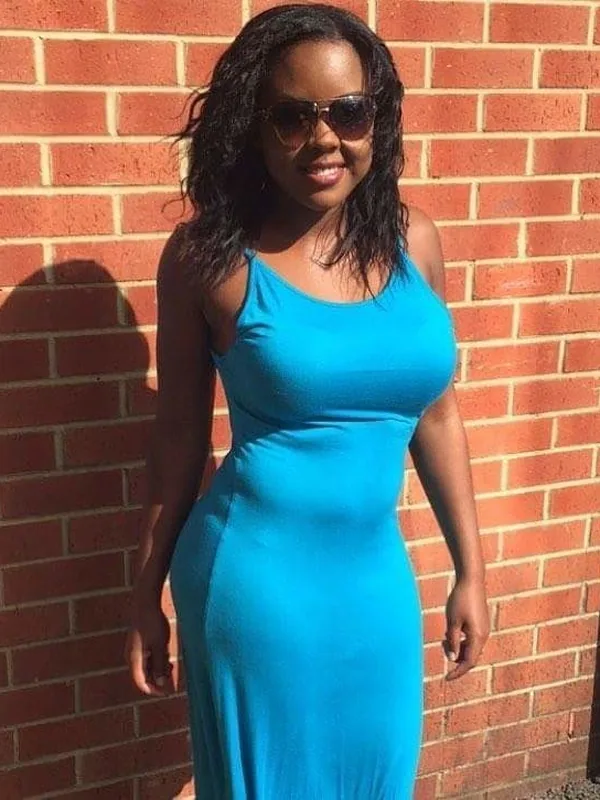 Femme souriante en robe bleue et lunettes de soleil devant un mur de briques rouges.
