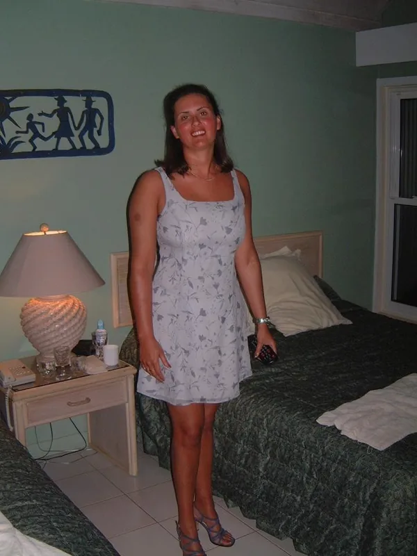 Femme souriante en robe blanche à motifs, debout dans une chambre avec un lit et une lampe de chevet allumée.