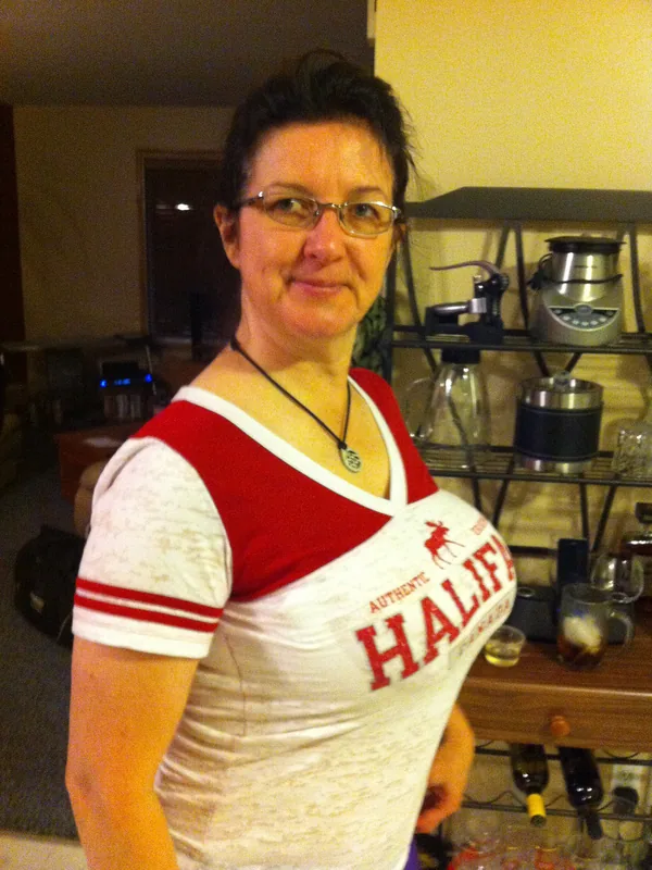 Femme aux cheveux courts et lunettes, en t-shirt blanc et rouge, dans un intérieur avec étagère de cuisine.