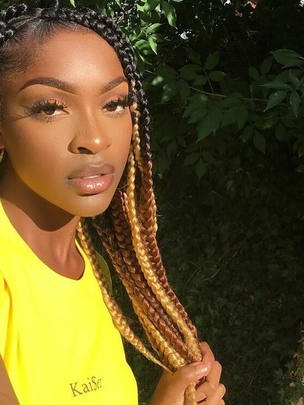 Portrait d'une femme aux cheveux tressés multicolores portant un t-shirt jaune, devant un arrière-plan végétal.