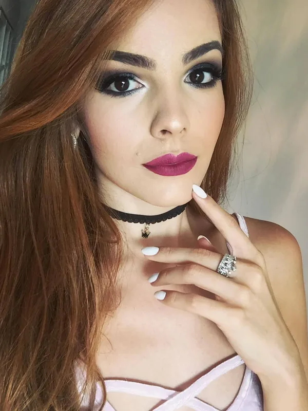 Portrait en gros plan d'une femme aux cheveux bruns, maquillée, portant un collier choker et une tenue rose pâle, la main près du menton.