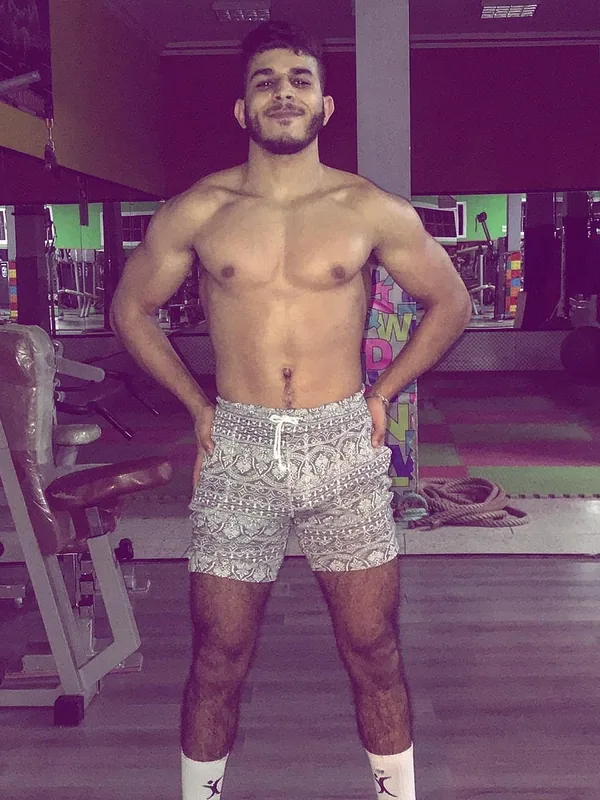 Homme torse nu en shorts à motifs, posant dans une salle de gym avec les mains sur les hanches.