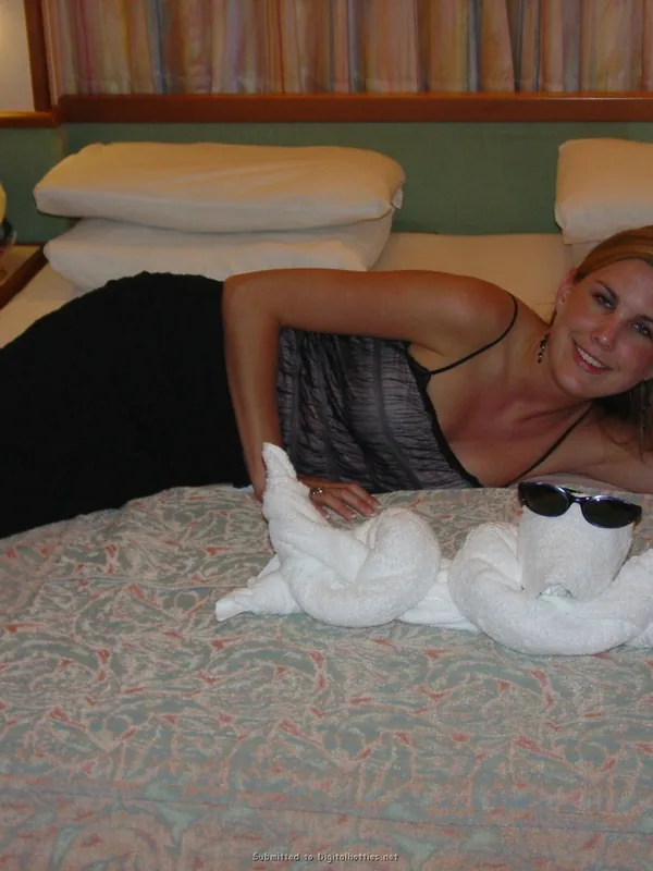 Femme allongée sur un lit avec des serviettes pliées en forme d'animaux, souriant vers l'objectif dans une chambre lumineuse.