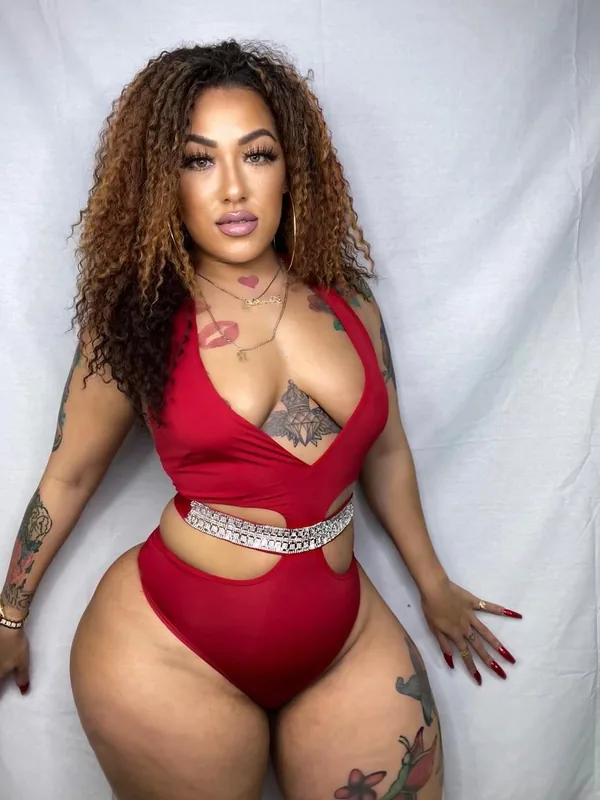 Femme aux cheveux bouclés portant un body rouge, tatouages visibles, regardant l'objectif devant un fond blanc.