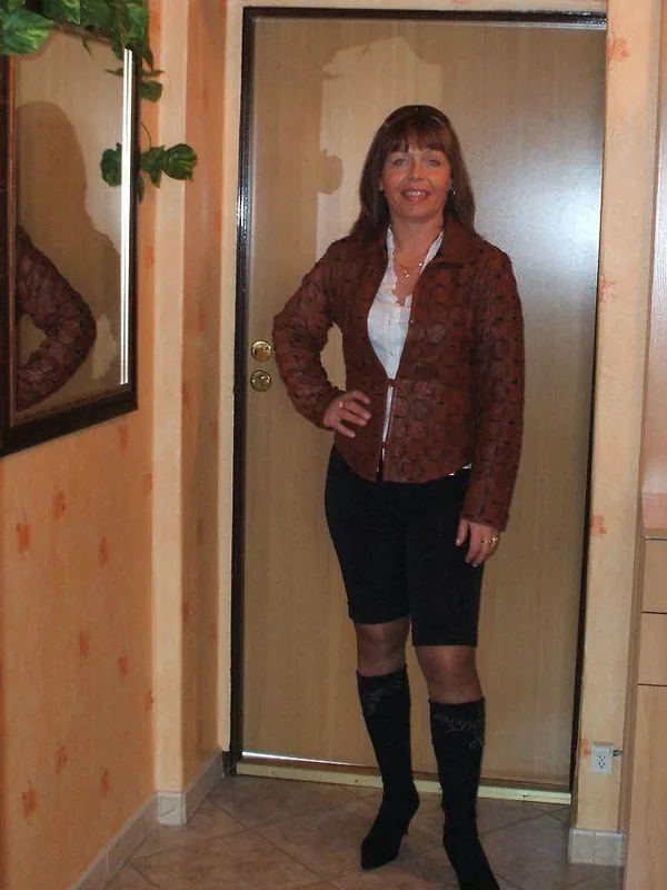 Femme debout dans un couloir, vêtue d'une veste marron, d'une jupe noire et de bottes hautes, souriant vers l'objectif.