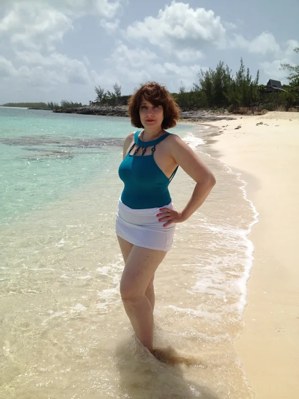 Femme en maillot de bain bleu et jupe blanche debout dans l'eau claire d'une plage tropicale.