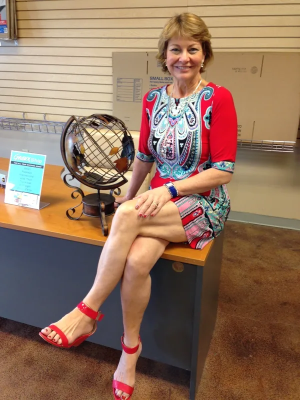 Femme blonde en robe rouge à motifs, assise sur un comptoir dans un magasin.
