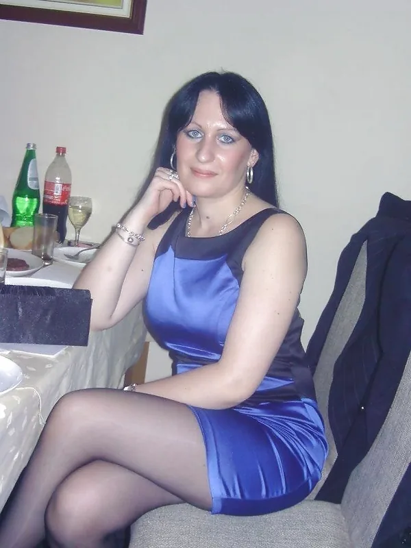 Femme aux cheveux noirs portant une robe bleue satinée, assise à une table avec des boissons, regardant l'objectif.