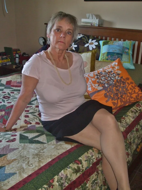 Femme aux cheveux courts gris assise sur un lit avec couverture patchwork, portant un t-shirt rose et une jupe noire, regardant l'objectif.