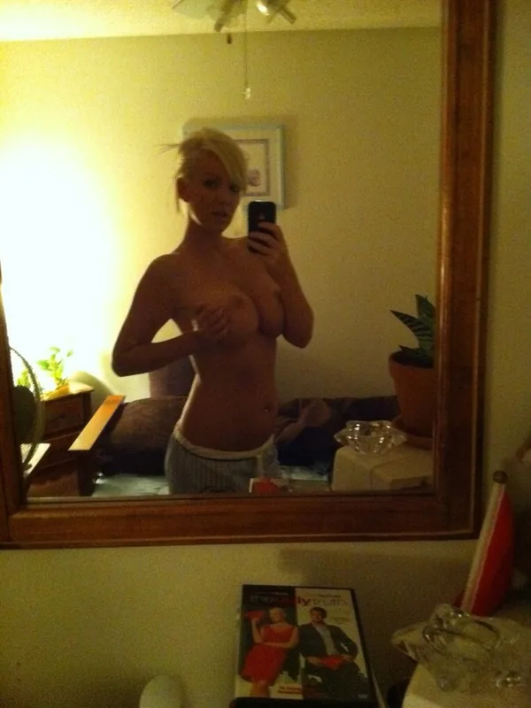 Femme blonde nue du torse, prenant un selfie dans un miroir, portant un short gris, dans une chambre éclairée par une lumière chaude.