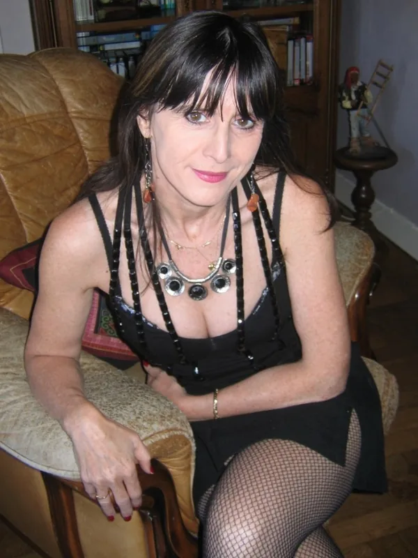 Femme assise sur un canapé en cuir, vêtue d'une tenue noire avec collants résille, regardant l'objectif dans un intérieur éclairé.
