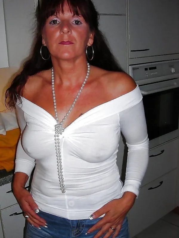Femme en haut blanc décolleté et jean bleu, debout dans une cuisine, regardant l'objectif.