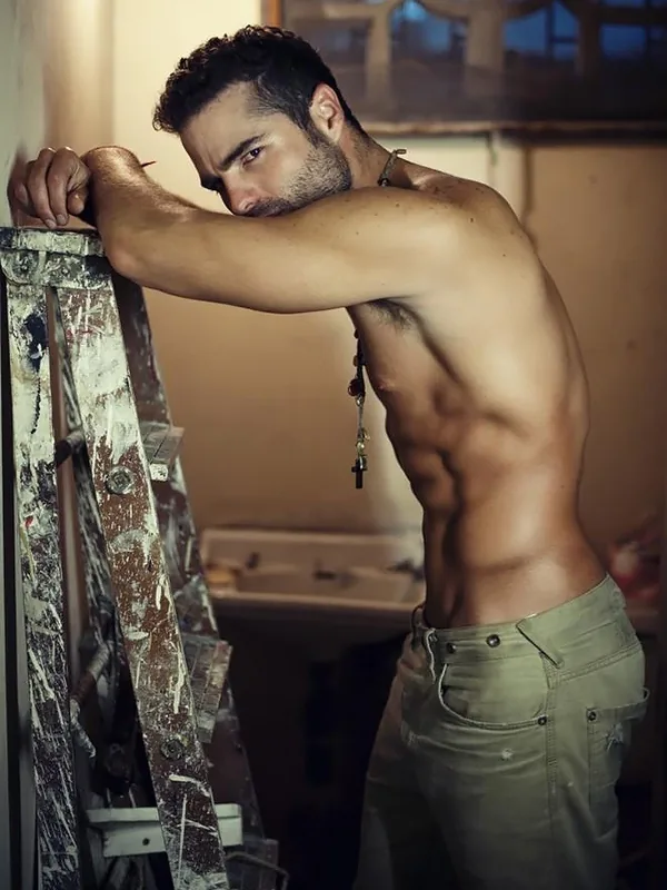 Homme torse nu, barbu, portant un collier avec croix et jeans, appuyé sur un échafaudage en bois dans un atelier.