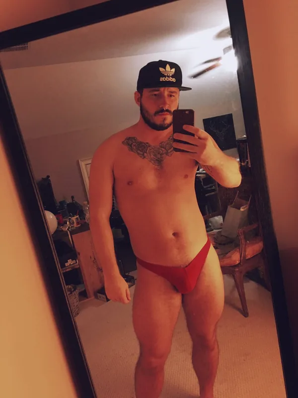 Homme torse nu en slip rouge et casquette Adidas prenant un selfie devant un miroir.