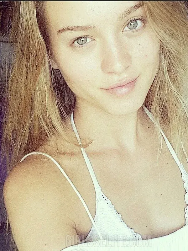 Portrait en gros plan d'une jeune femme aux cheveux blonds portant un haut blanc à bretelles, regardant l'objectif dans une chambre.