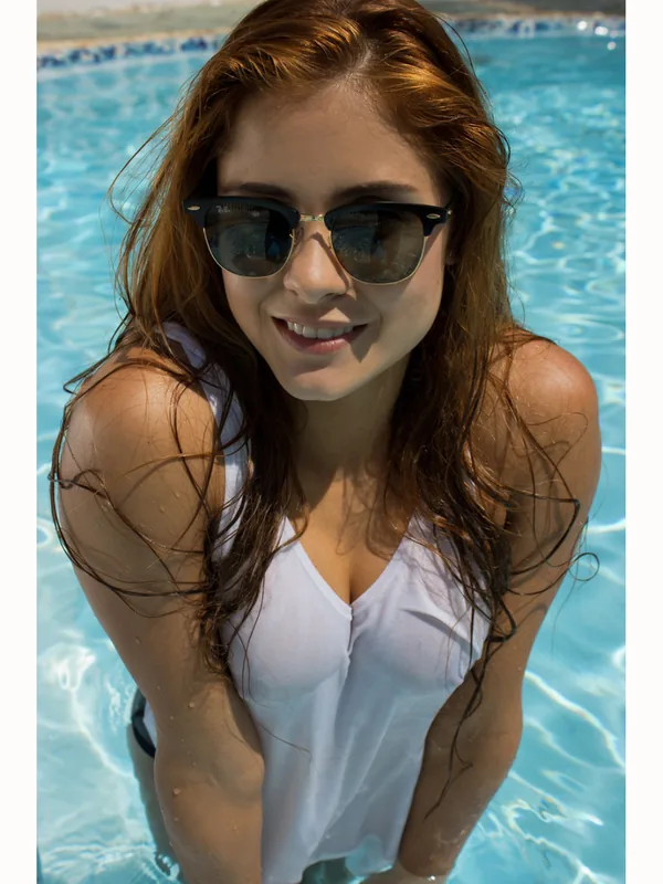 Femme souriante avec lunettes de soleil, cheveux mouillés, portant un haut blanc, dans une piscine sous la lumière du soleil.