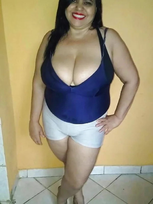 Femme souriante en haut bleu et short blanc, debout devant un mur jaune.