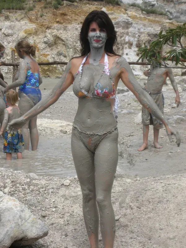 Femme couverte de boue en maillot de bain, debout dans un site naturel avec d'autres personnes autour.