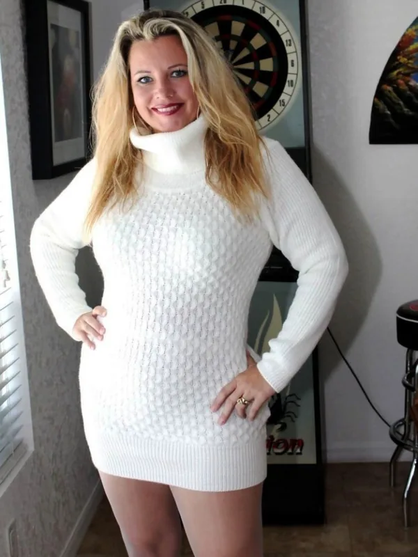 Femme blonde en pull blanc à col roulé, souriante, posant devant un jeu de fléchettes dans une pièce intérieure.