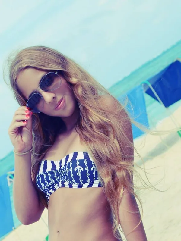 Femme aux cheveux blonds portant un haut de maillot de bain bleu et blanc et des lunettes de soleil sur une plage ensoleillée.