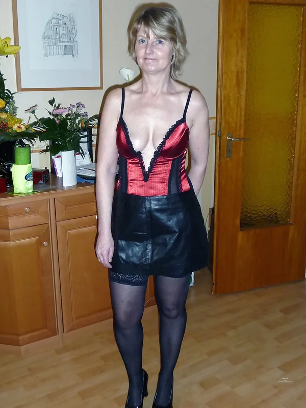 Femme debout en tenue rouge et noire, jupe courte, collants et talons, dans un intérieur avec meubles en bois.