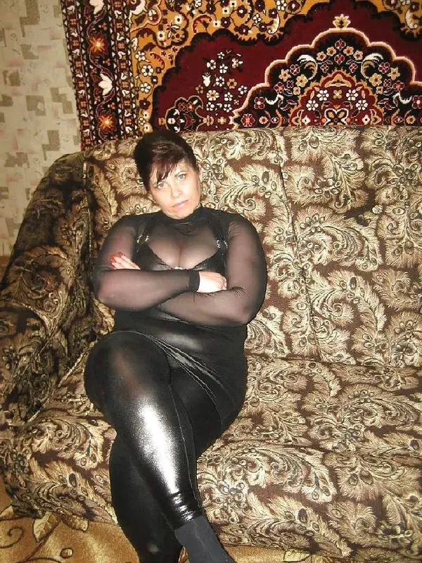 Femme assise sur un canapé à motifs, vêtue d'une tenue noire transparente et brillante, regardant l'objectif.