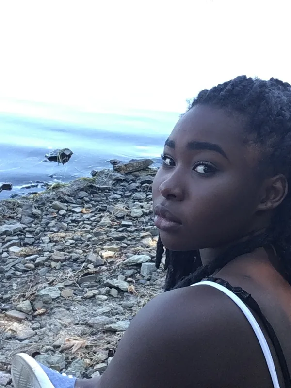 Jeune femme aux tresses noires assise sur un rivage rocheux, regardant l'objectif, avec la mer en arrière-plan.