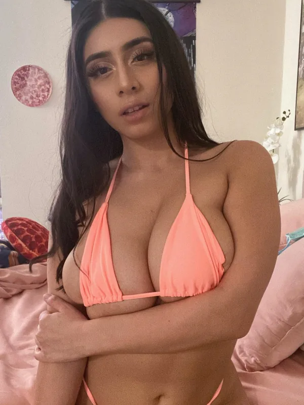 Femme aux cheveux noirs portant un bikini rose pêche, assise sur un lit rose, regardant l'objectif.