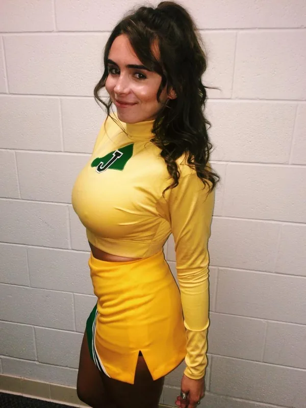 Femme en tenue jaune de cheerleader avec une lettre J verte, posant devant un mur blanc.