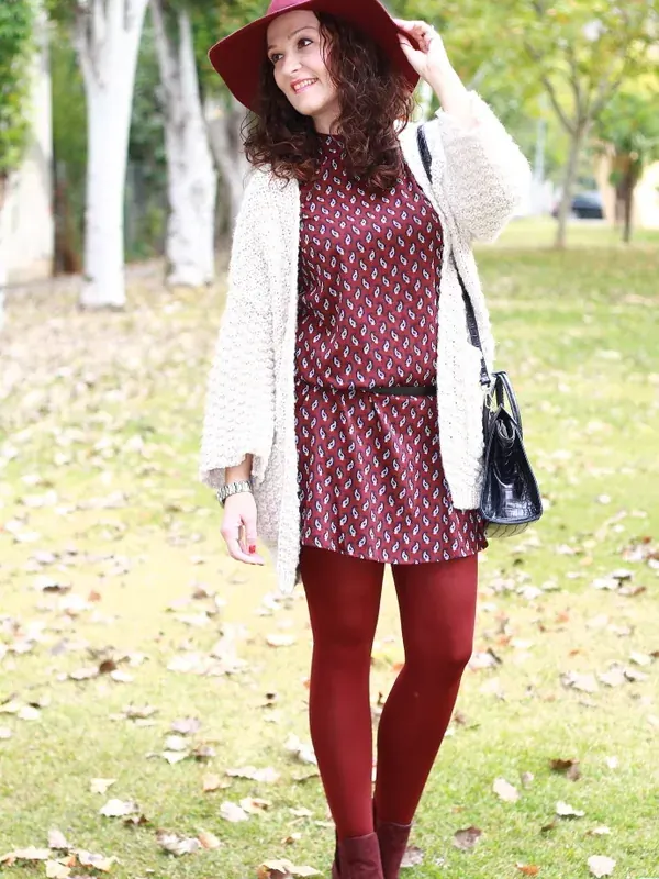 Femme souriante en tenue bordeaux et blanche, portant un chapeau, debout sur une pelouse en automne.