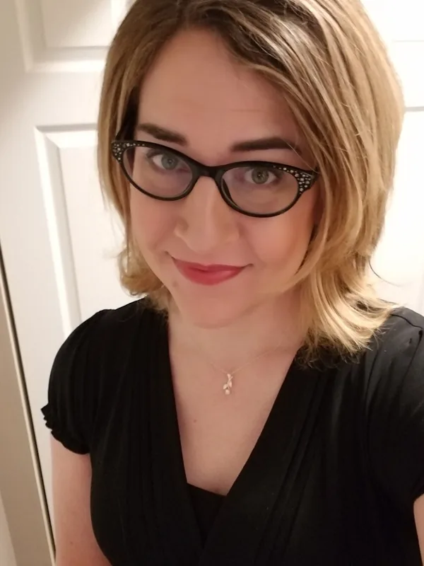Portrait en buste d'une femme aux cheveux blonds, lunettes noires, robe noire, regardant l'objectif.