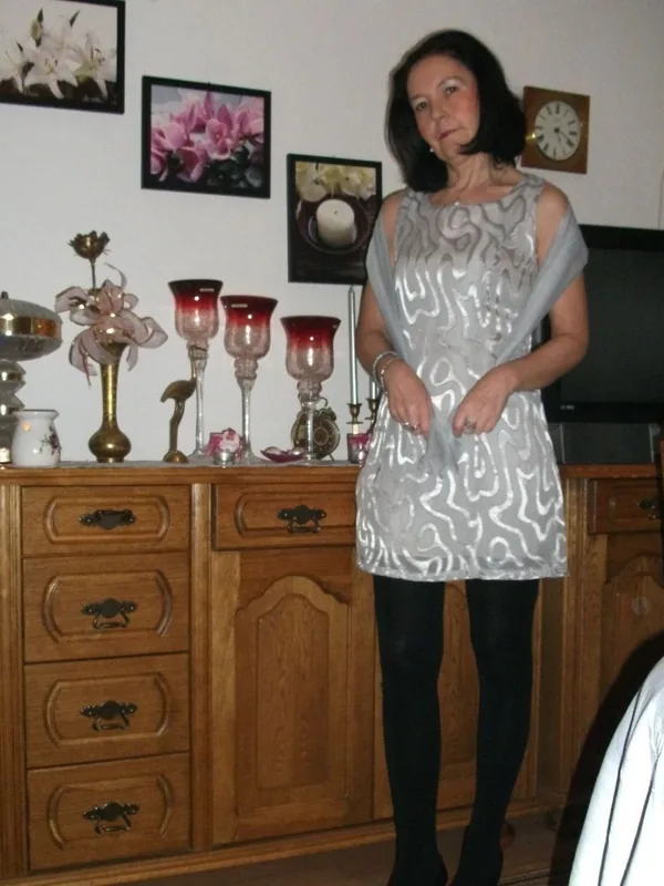 Femme debout dans une chambre, devant une commode en bois, portant une robe grise et des collants noirs.