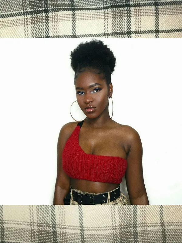 Portrait en buste d'une femme aux cheveux noirs en chignon, portant un haut rouge asymétrique et des boucles d'oreilles dorées.