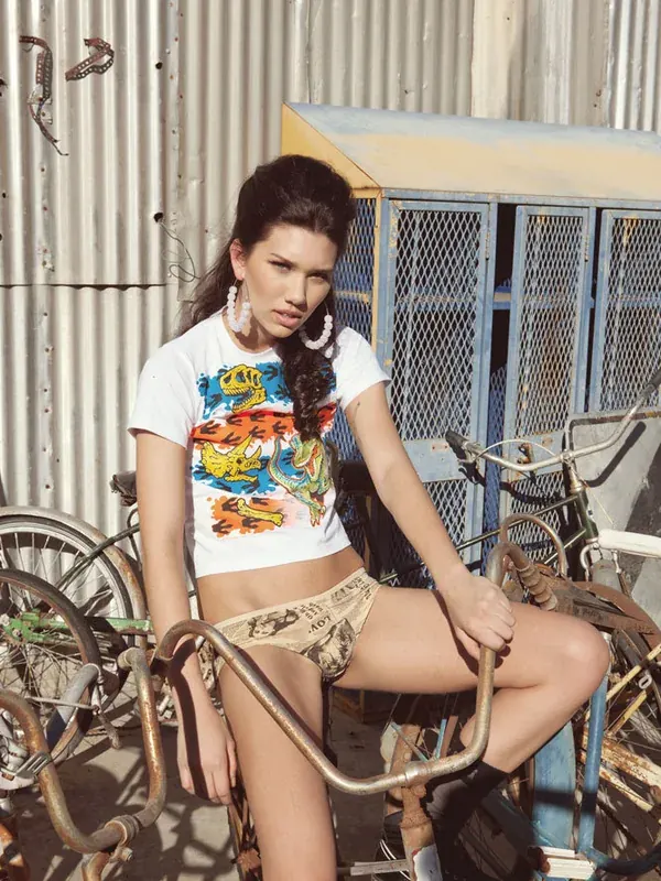 Femme assise sur un cadre de vélo rouillé, portant un t-shirt à dinosaures et des sous-vêtements, devant une clôture en tôle ondulée.