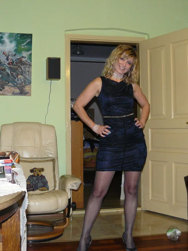 Femme en robe noire souriante, debout dans un salon, devant une porte ouverte, entourée de meubles et d'objets personnels.
