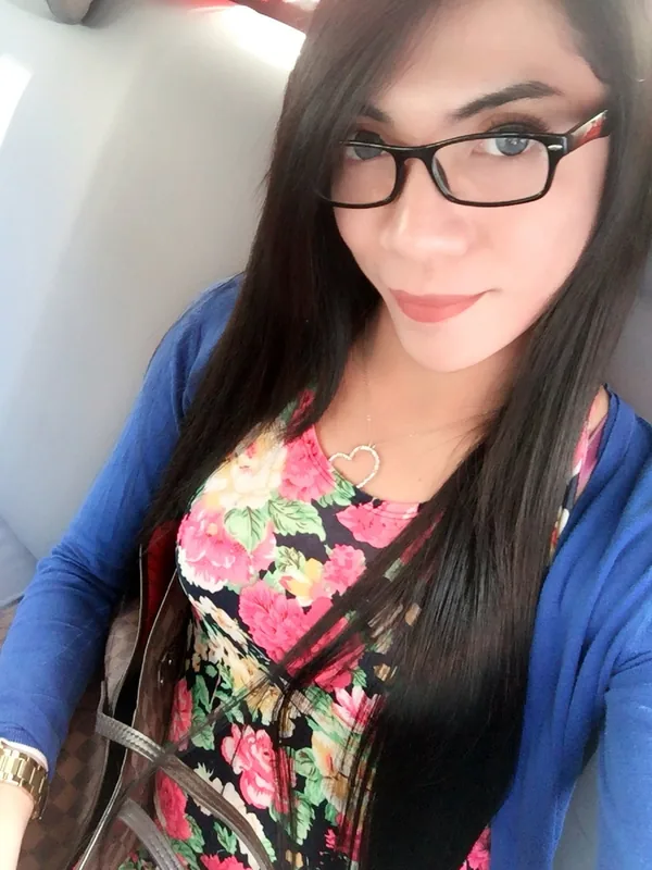 Portrait d'une femme aux cheveux longs noirs portant des lunettes, un haut floral et une veste bleue, assise dans un véhicule.