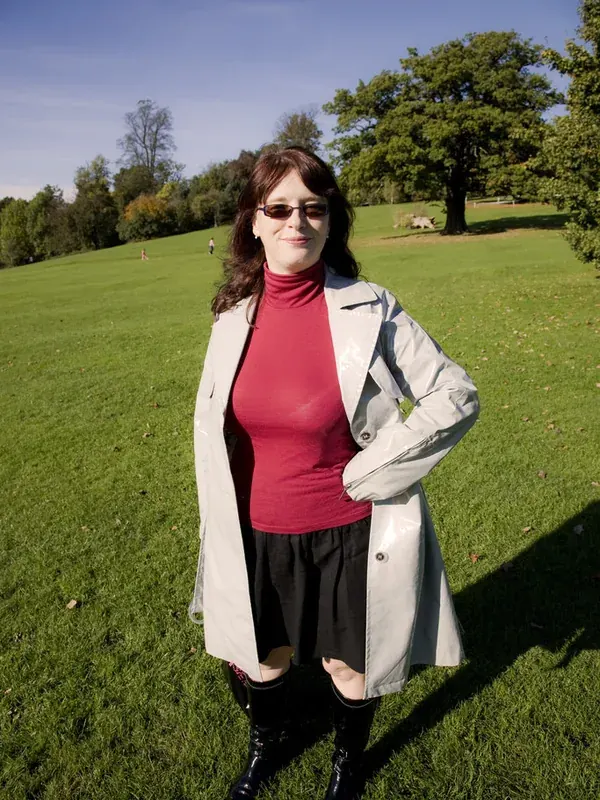 Femme debout sur une pelouse ensoleillée, portant un manteau gris, un haut rouge, une jupe noire et des bottes, avec des lunettes de soleil.