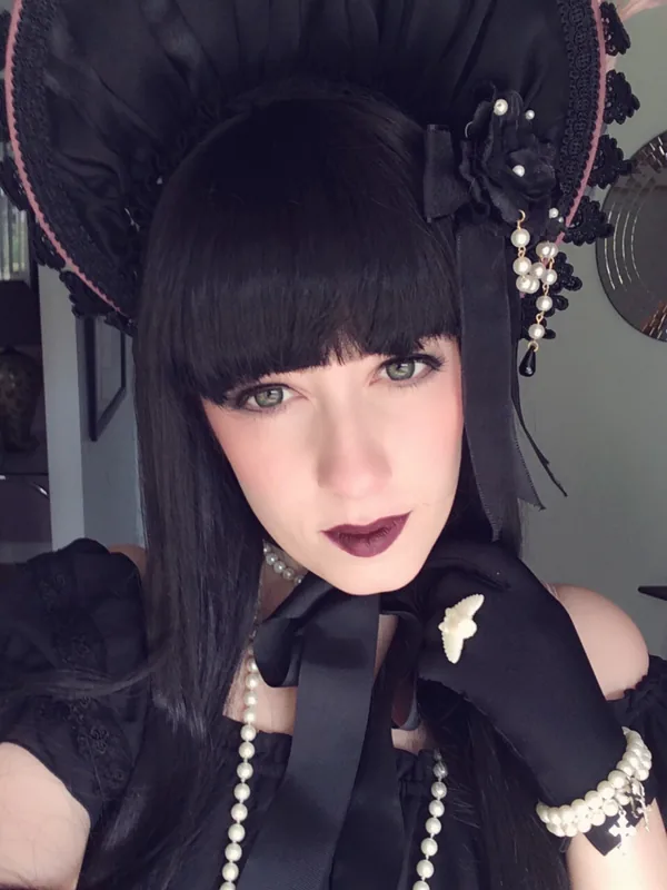 Femme aux cheveux noirs portant un chapeau noir orné de perles, une robe gothique, des gants et un collier de perles, regardant l'objectif.