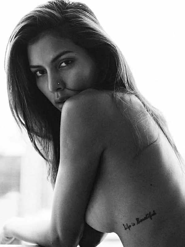 Portrait en noir et blanc d'une femme aux cheveux longs, nue du torse, regardant l'objectif avec un tatouage visible sur le dos.