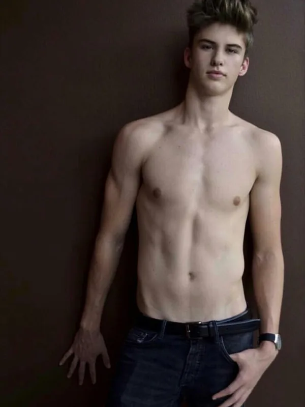 Jeune homme torse nu, portant un jean et une ceinture, appuyé contre un mur sombre, regardant l'objectif.