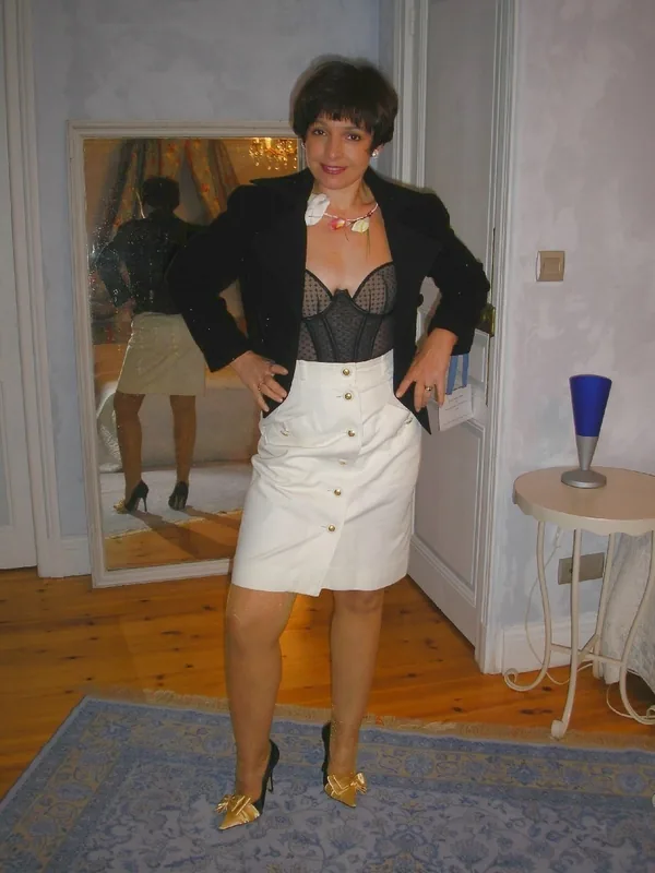 Femme aux cheveux courts portant une lingerie noire, une veste noire et une jupe blanche, posant devant un miroir dans une pièce éclairée.
