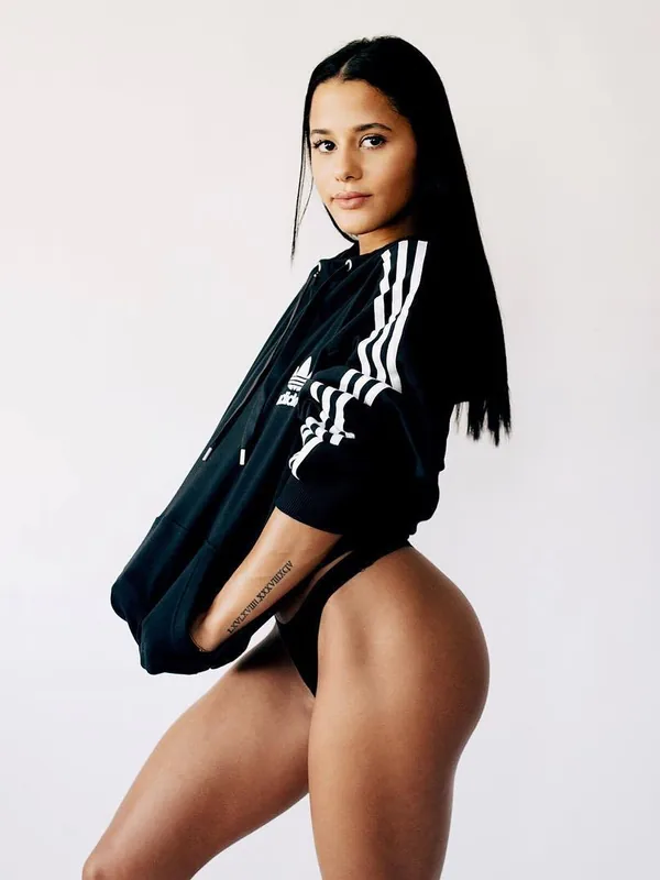 Femme aux cheveux noirs portant une veste de sport noire ouverte, posant de profil sur fond blanc.