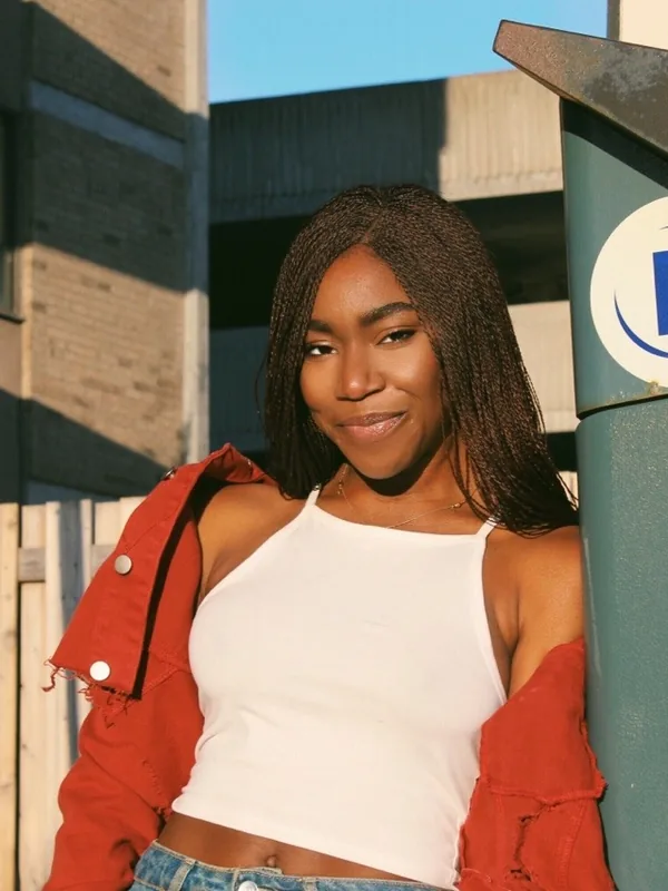 Jeune femme aux cheveux tressés portant un haut blanc et une veste rouge, appuyée contre un distributeur de stationnement en plein jour.