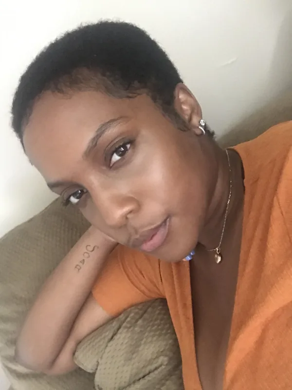 Portrait d'une femme aux cheveux courts portant un haut orange, posant sur un coussin beige avec un tatouage au poignet.