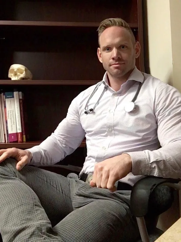 Homme en chemise blanche et pantalon gris assis dans un fauteuil de bureau devant une étagère avec livres et crâne.