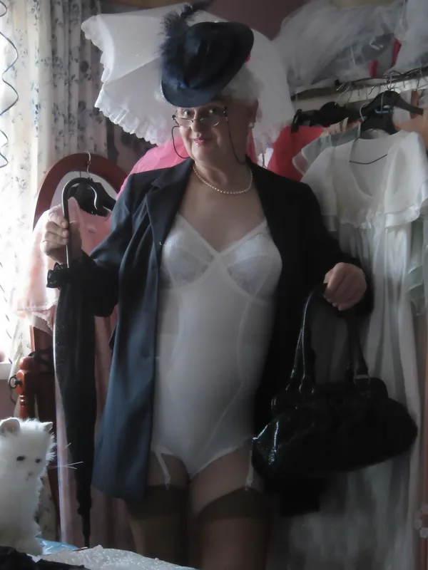 Femme portant un chapeau noir, une veste, un body blanc et des collants, tenant un parapluie et un sac, avec un chat blanc à ses pieds.