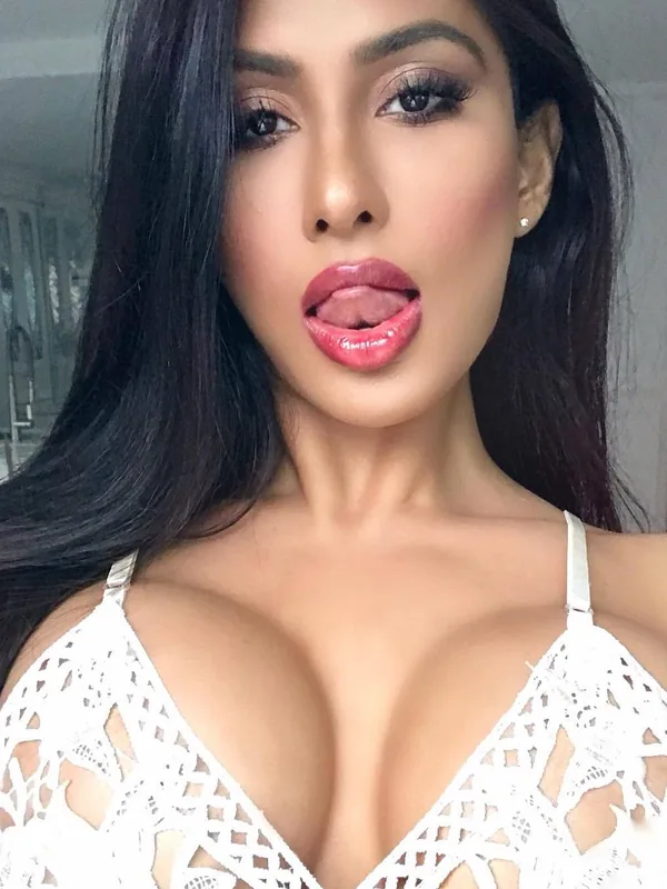 Portrait d'une femme aux cheveux noirs portant un haut blanc en dentelle, regardant l'objectif avec la langue légèrement sortie.