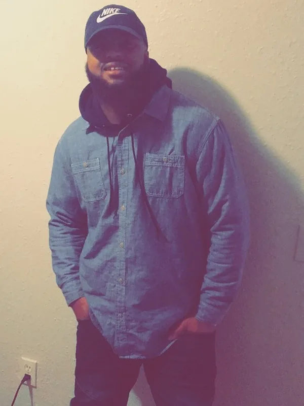 Homme barbu en casquette Nike et chemise en jean, debout contre un mur clair, mains dans les poches.